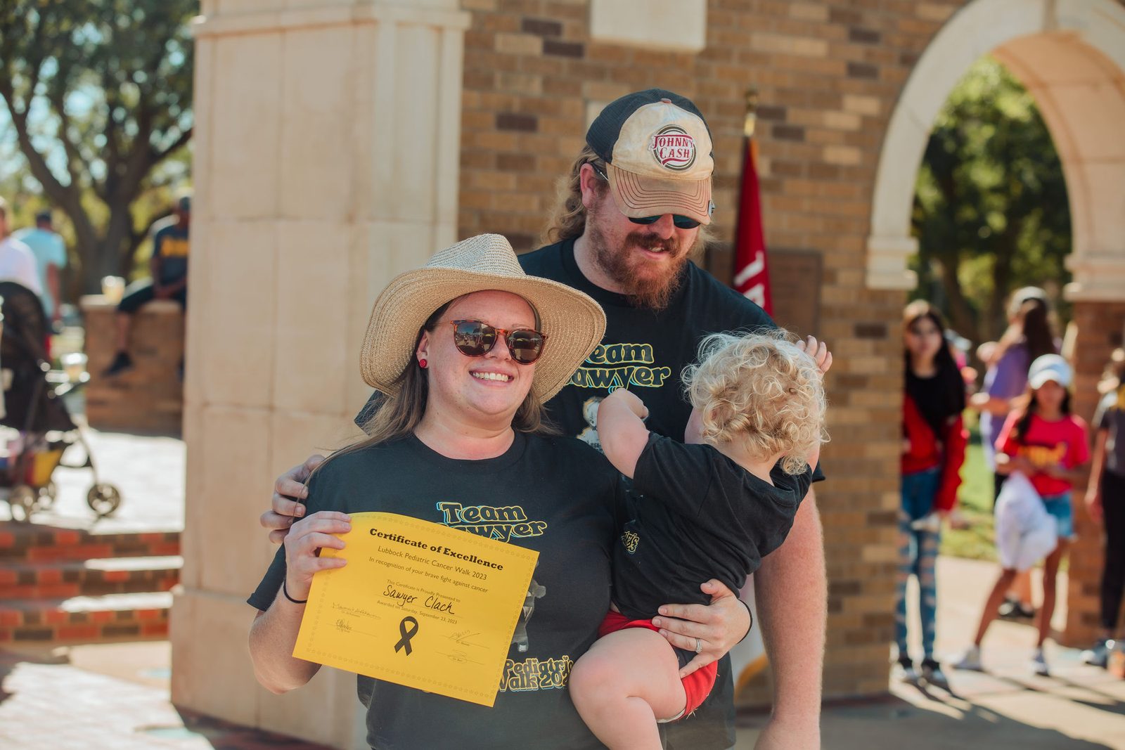 A family in matching team shirts holds a gold LPCA certificate of excellence at the Pediatric Cancer Walk.